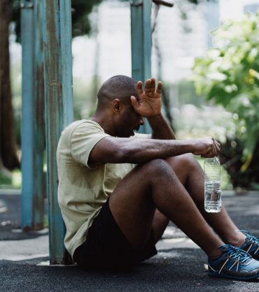 Exhausted man sitting against a pole while holding a water bottle