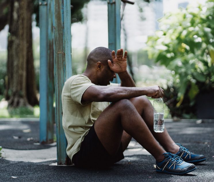 Exhausted man sitting against a pole while holding a water bottle